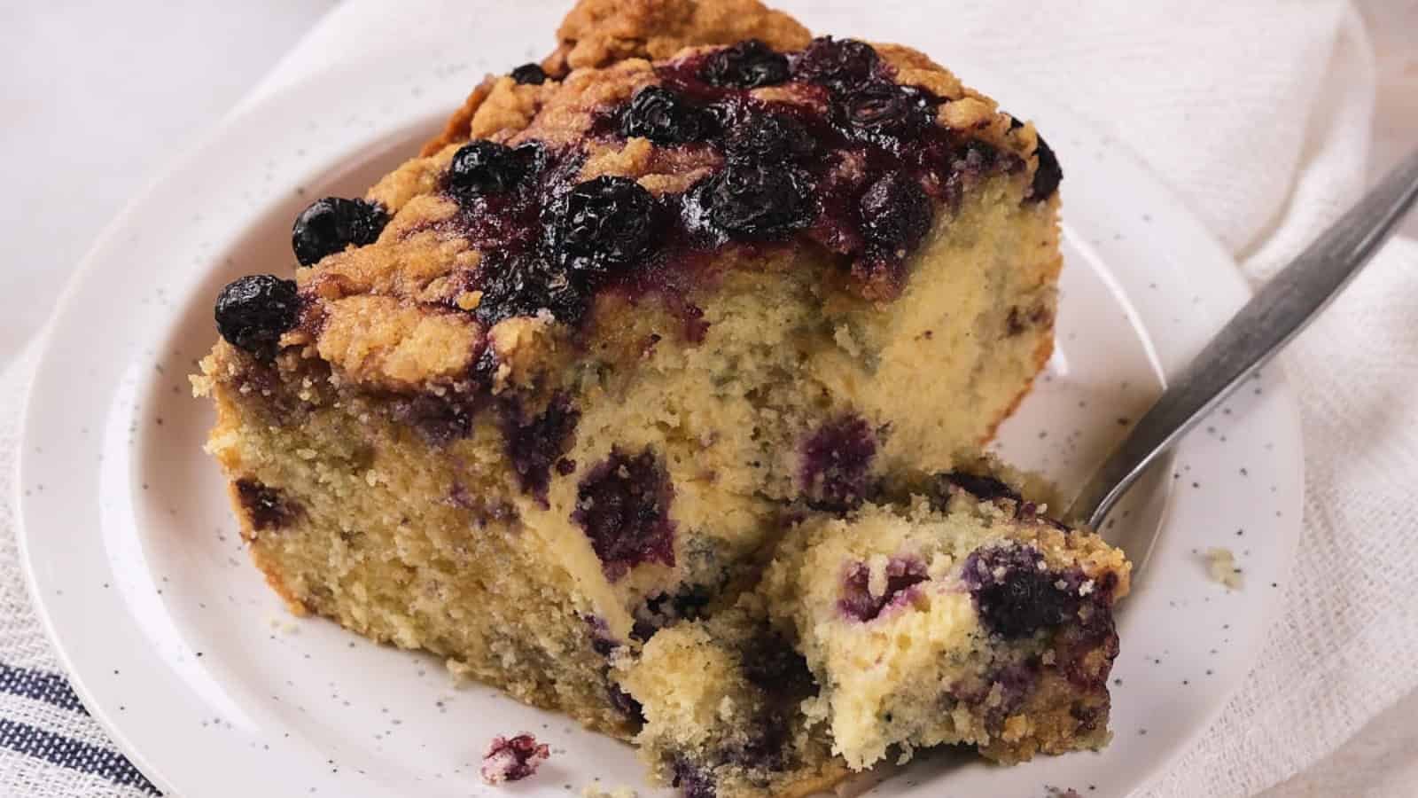 A slice of blueberry coffee cake on a white plate with a fork, showing a crumbly topping and visible blueberries throughout the cake.