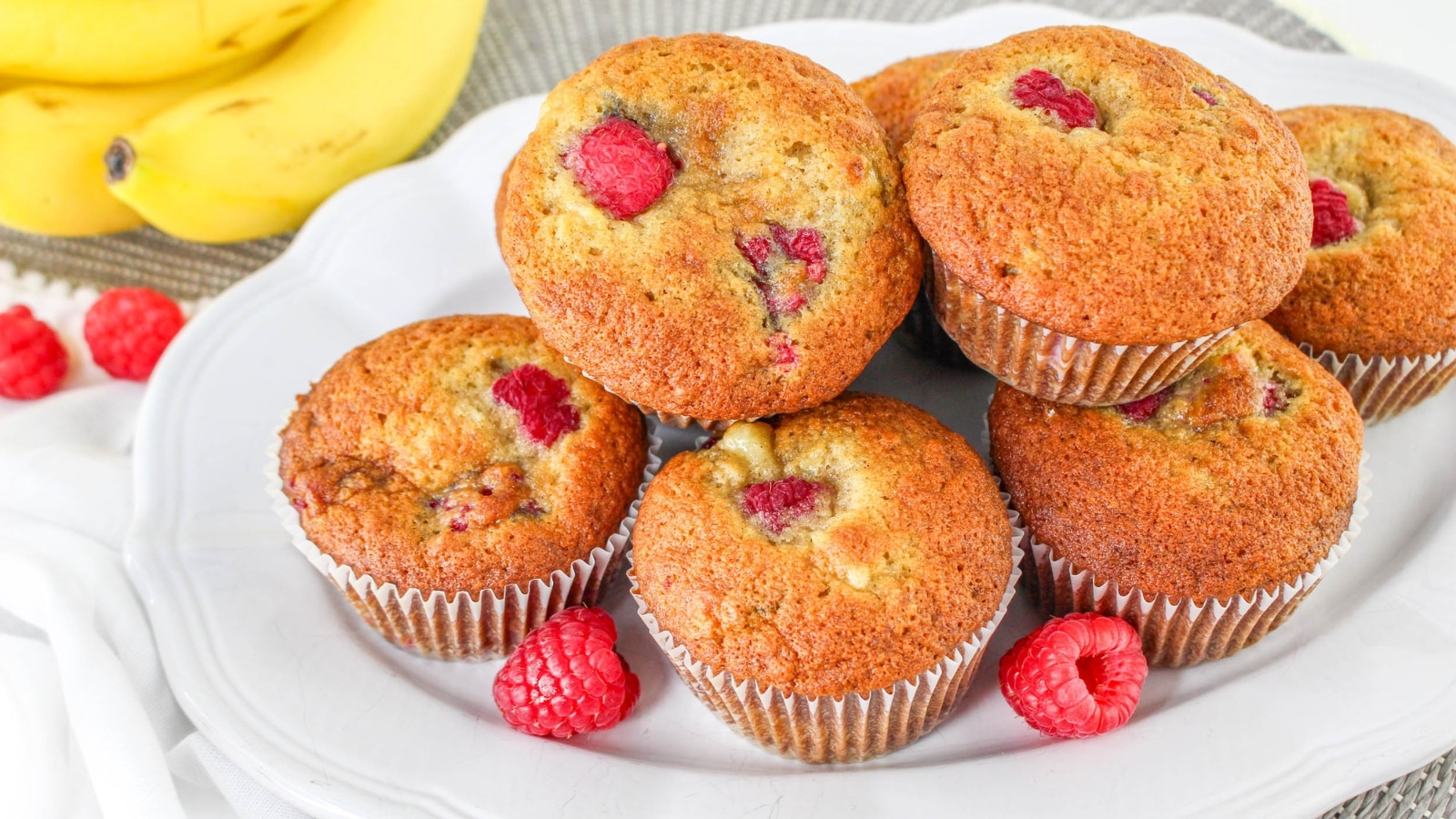 A plate of banana raspberry muffins is arranged with fresh raspberries, with bananas visible in the background.