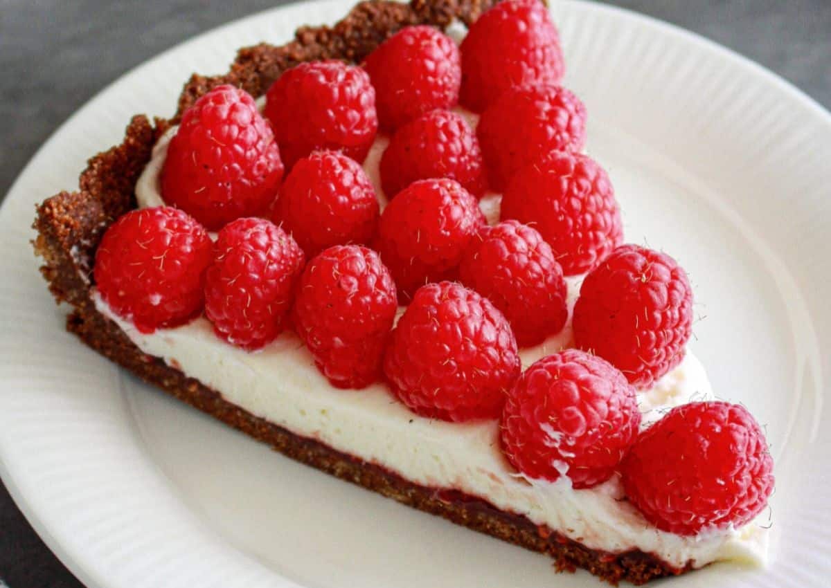 A slice of tart with a chocolate crust, creamy filling, and fresh raspberries arranged on top, served on a white plate.