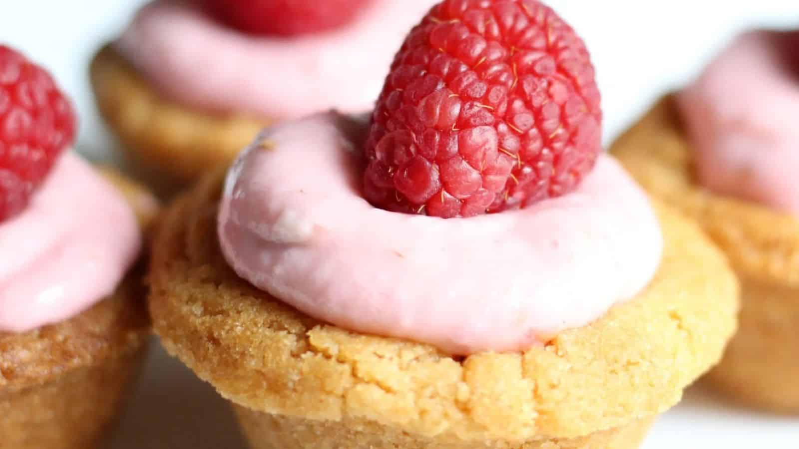 Close-up of a mini tart with a golden crust, pink cream filling, and a fresh raspberry on top.