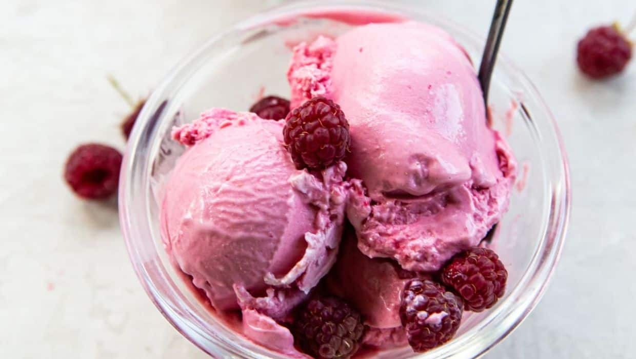 A glass bowl filled with three scoops of pink raspberry ice cream, garnished with whole raspberries and a spoon.