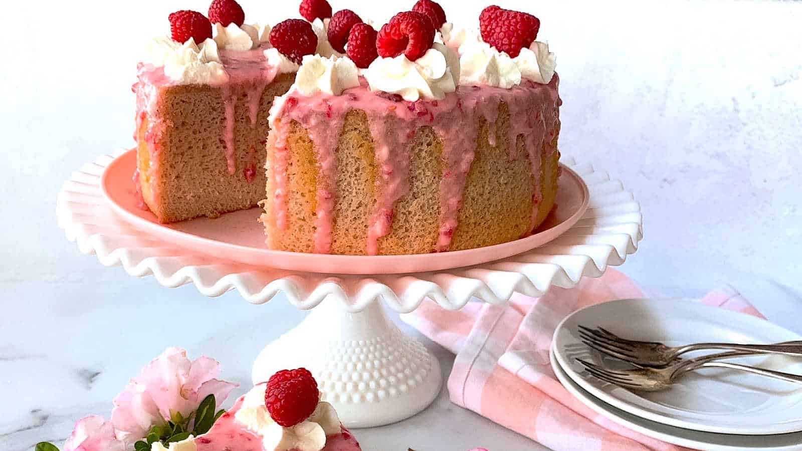 A sponge cake with pink glaze, whipped cream, and raspberries on top sits on a cake stand; a slice is missing. Plates, forks, and a pink napkin are nearby.