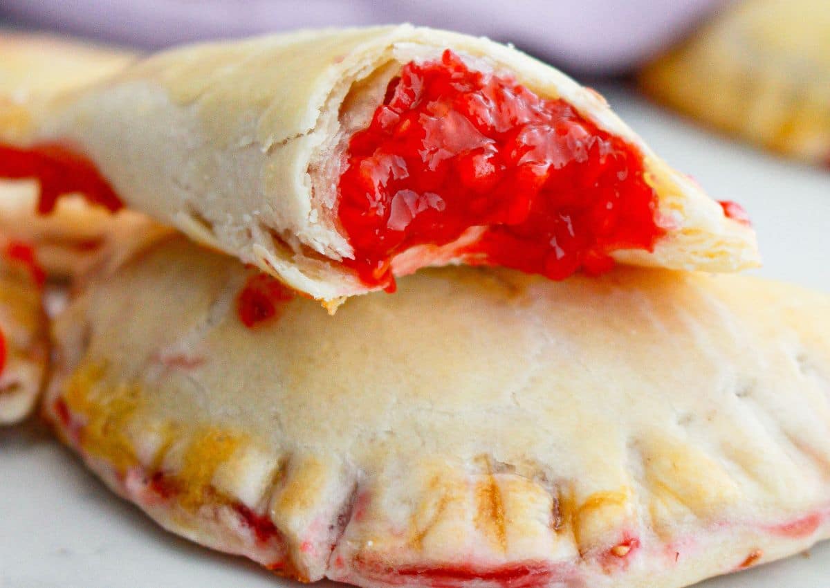 A close-up of a baked hand pie with a golden crust, broken open to reveal a bright red cherry filling inside.
