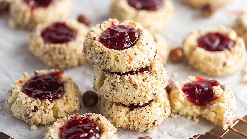 A stack of thumbprint cookies filled with red jam and coated with chopped nuts, arranged on parchment paper with more cookies scattered around.