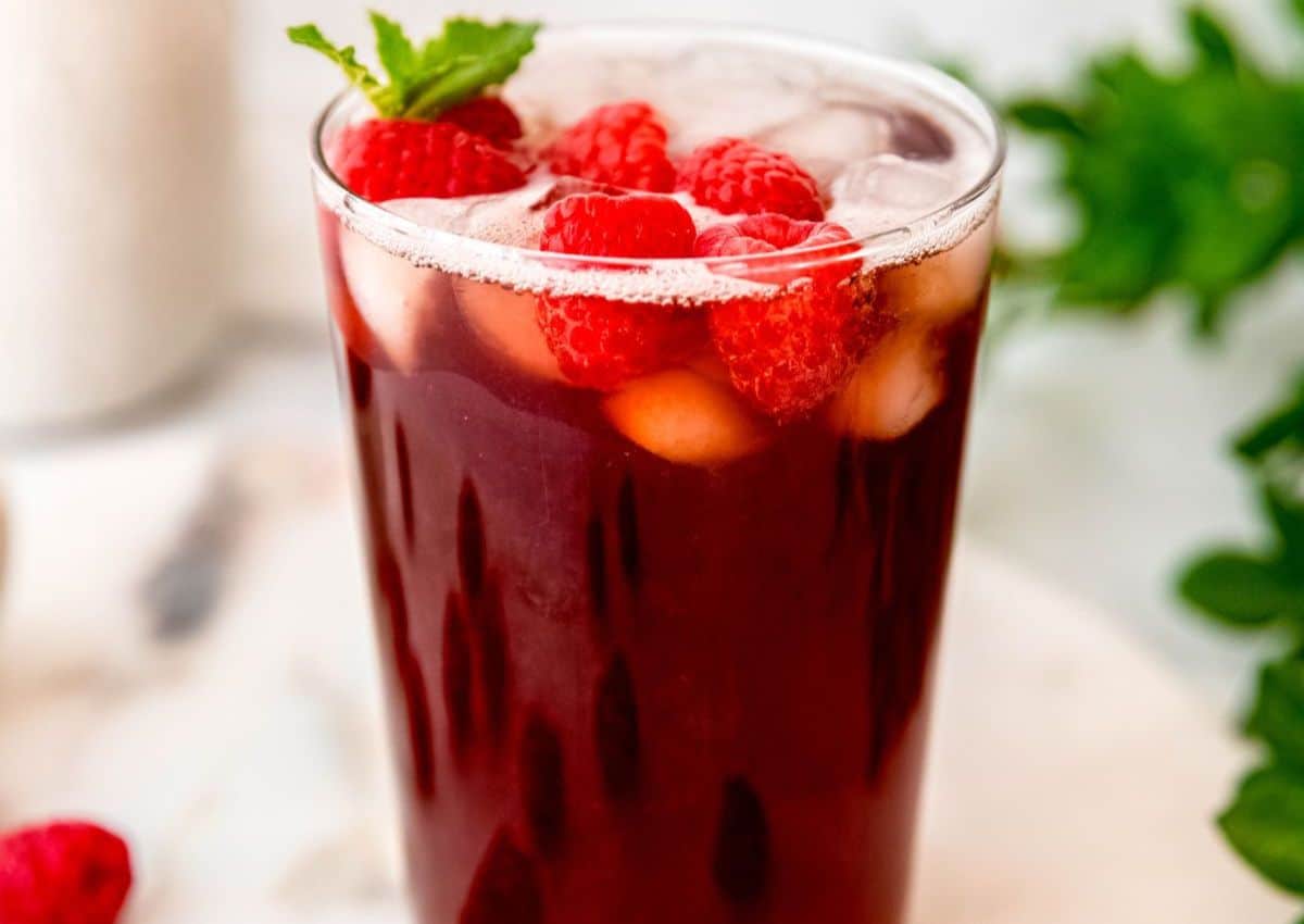 A glass of iced red beverage garnished with fresh raspberries and a mint sprig on a white surface.