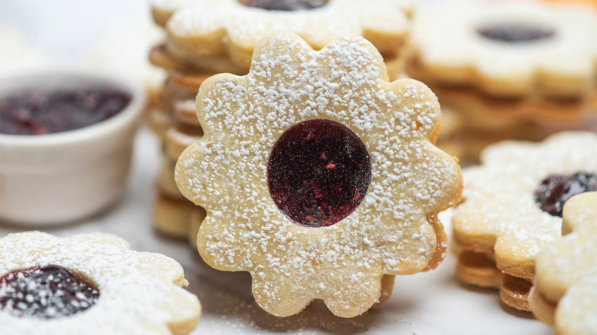 Round, flower-shaped sandwich cookies with a center of red jam and dusted with powdered sugar are stacked on a white surface. A small bowl of jam is in the background.