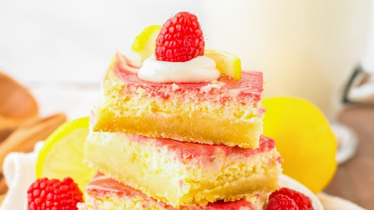 Three lemon raspberry dessert bars stacked, each topped with raspberry, lemon slice, and a dollop of cream, with more raspberries and lemon slices around them.
