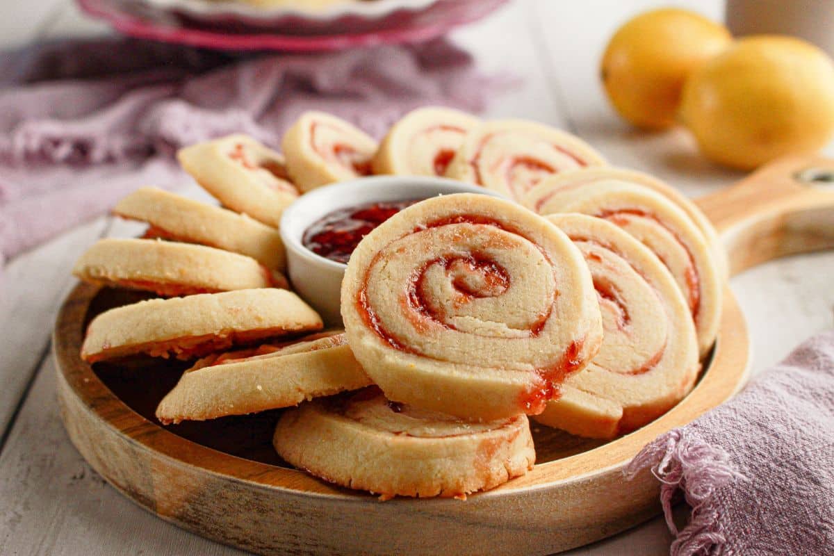 A wooden serving board with swirled jam cookies arranged around a small dish of jam, set on a table with a purple napkin and two yellow fruits in the background.