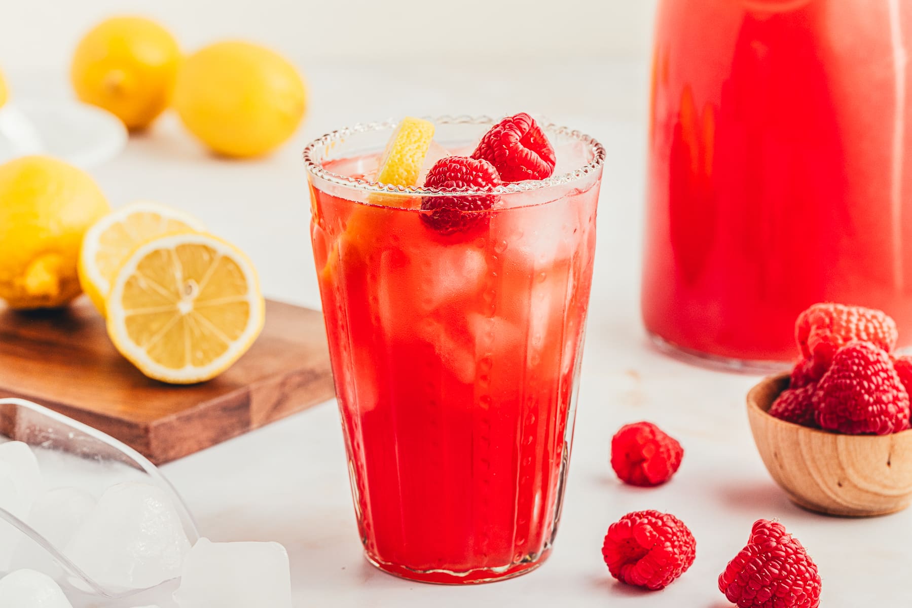 A glass of raspberry lemonade with ice, garnished with lemon slices and raspberries, sits on a white surface with more fruit and a pitcher in the background.