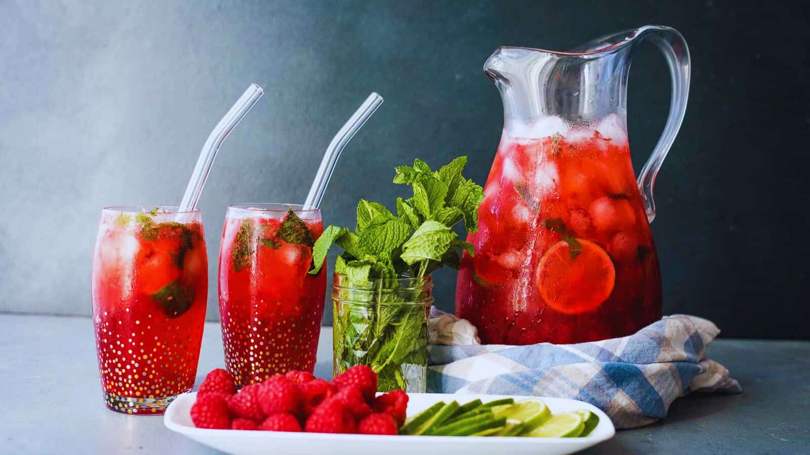 A pitcher and two glasses of red iced drink with mint and fruit, next to a jar of mint, fresh raspberries, lime slices, and cucumber on a white plate.