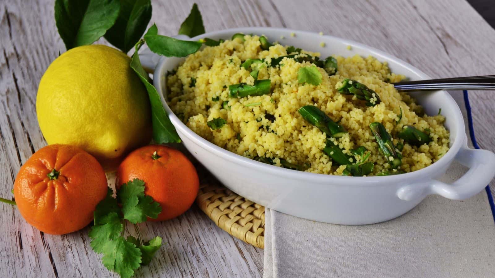 A white dish filled with couscous and green vegetables sits beside a lemon, two tangerines, and some cilantro on a wooden surface.