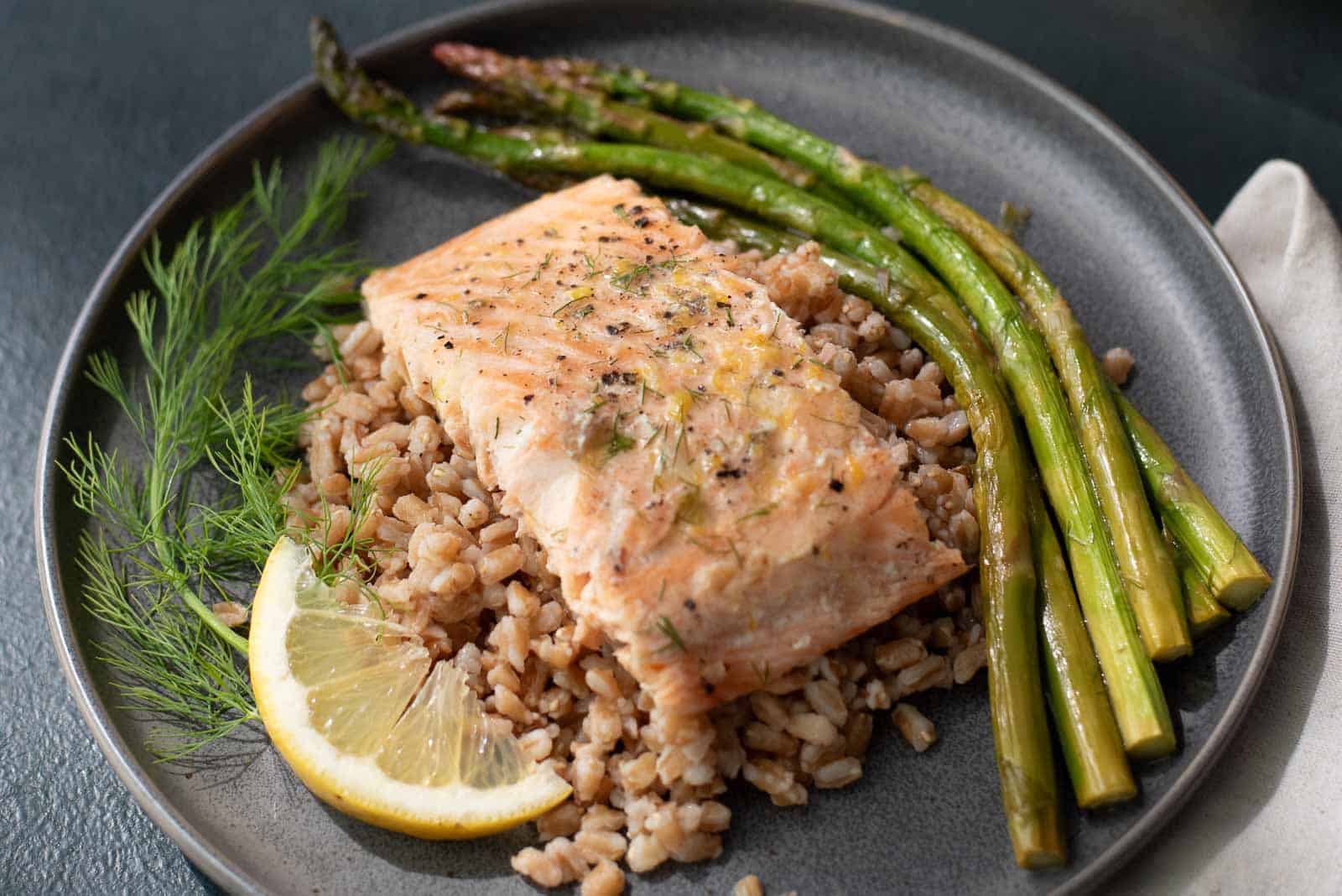A plate with a cooked salmon fillet on a bed of grains, served with roasted asparagus, a lemon slice, and a sprig of dill.