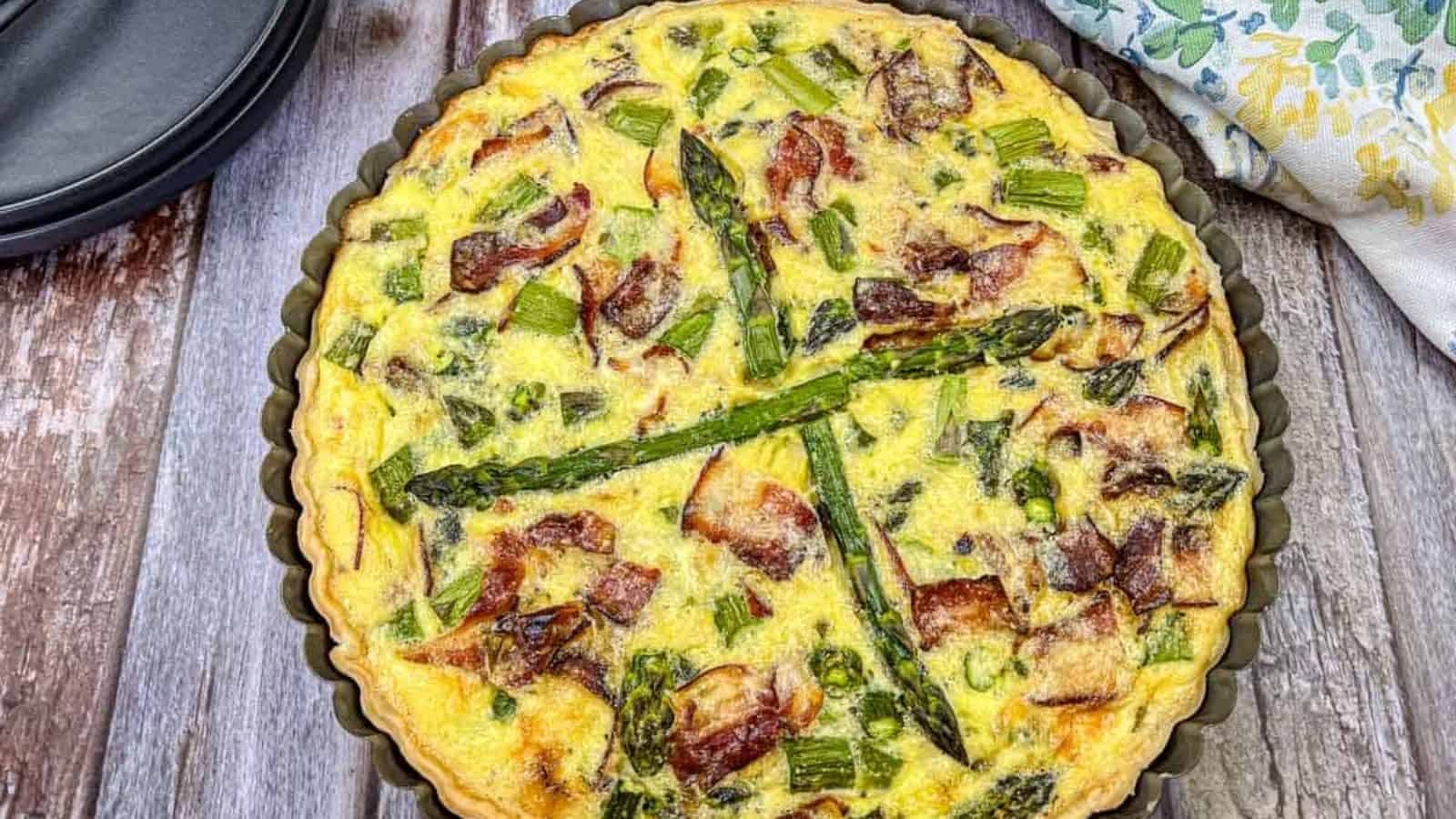 A baked quiche in a fluted pan, filled with asparagus, bacon, and other vegetables, sits on a wooden surface next to a patterned cloth.