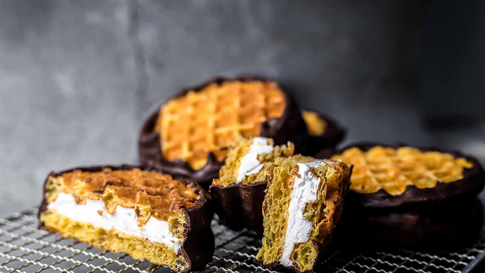 Chocolate-dipped waffle sandwiches filled with marshmallow cream are displayed on a cooling rack, with one sandwich cut in half to show the filling.