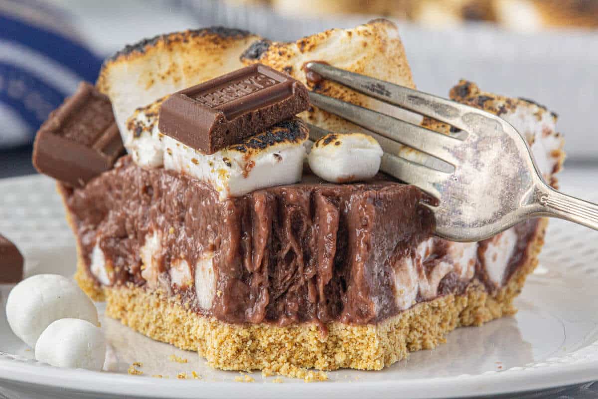 A slice of smores pie with a graham cracker crust, chocolate filling, toasted marshmallows, and chocolate pieces, being cut with a fork on a white plate.