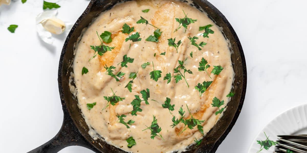 A cast iron skillet filled with creamy chicken in a white sauce, garnished with chopped parsley.