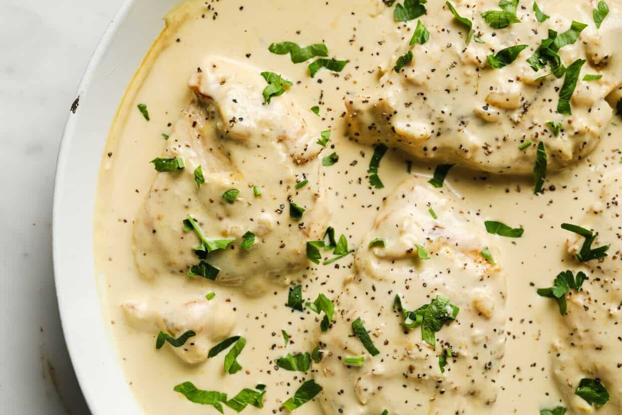 Chicken breasts covered in a creamy sauce, garnished with chopped parsley and black pepper, served in a white dish.