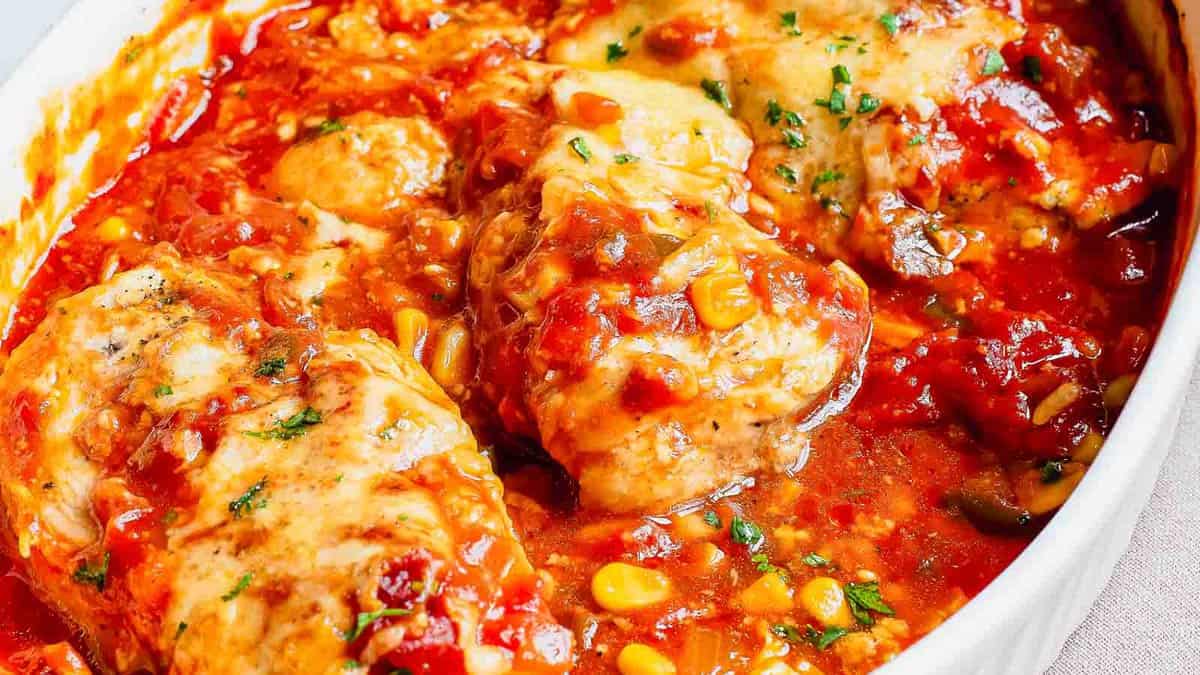 Baked chicken breasts in a tomato-based sauce with corn, chopped vegetables, and herbs in a white dish.