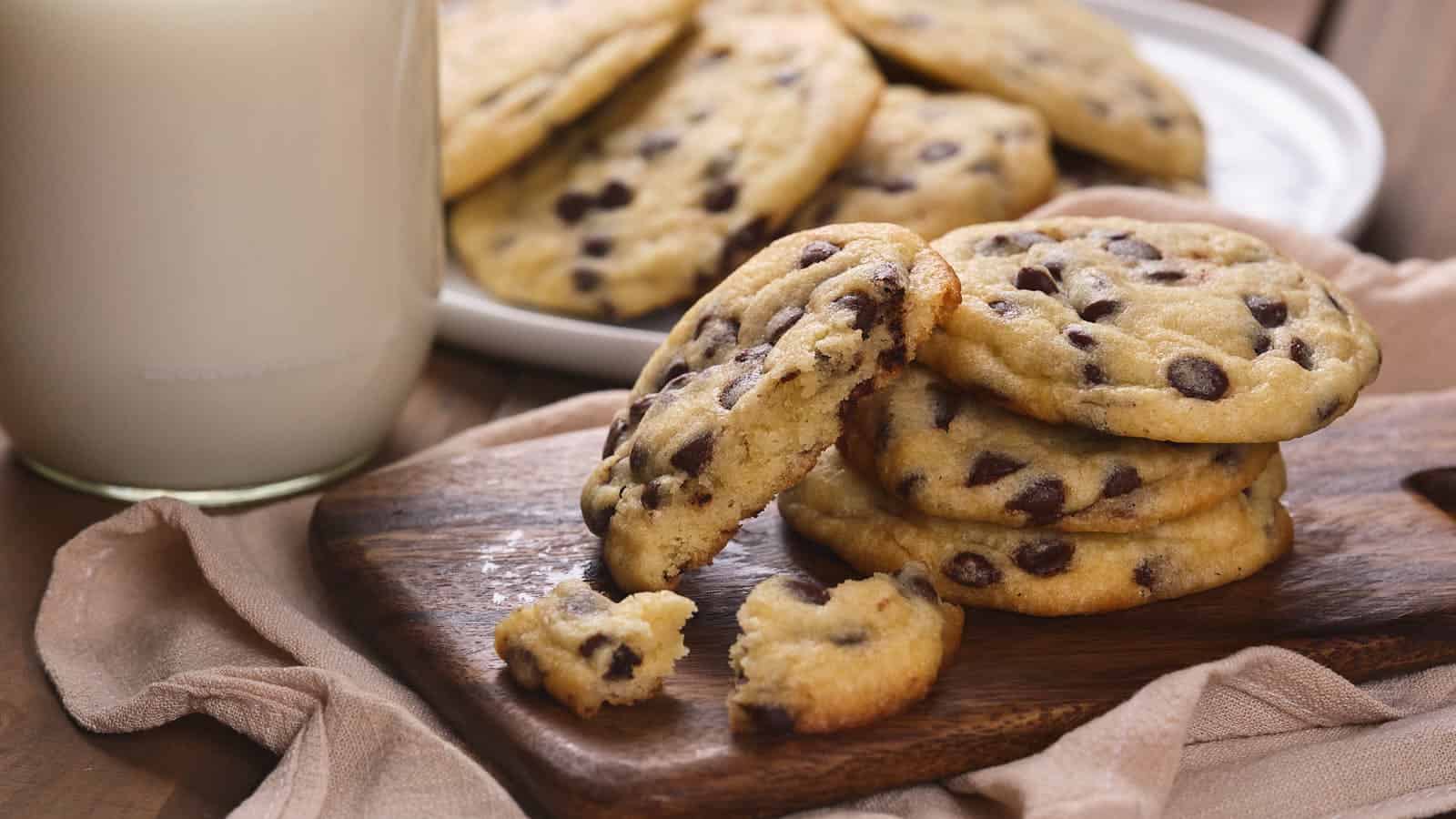 A stack of chocolate chip cookies sits on a wooden board next to a glass of milk, perfect for celebrating National Chocolate Chip Cookie Day recipes, with more cookies on a plate and one broken cookie in front.