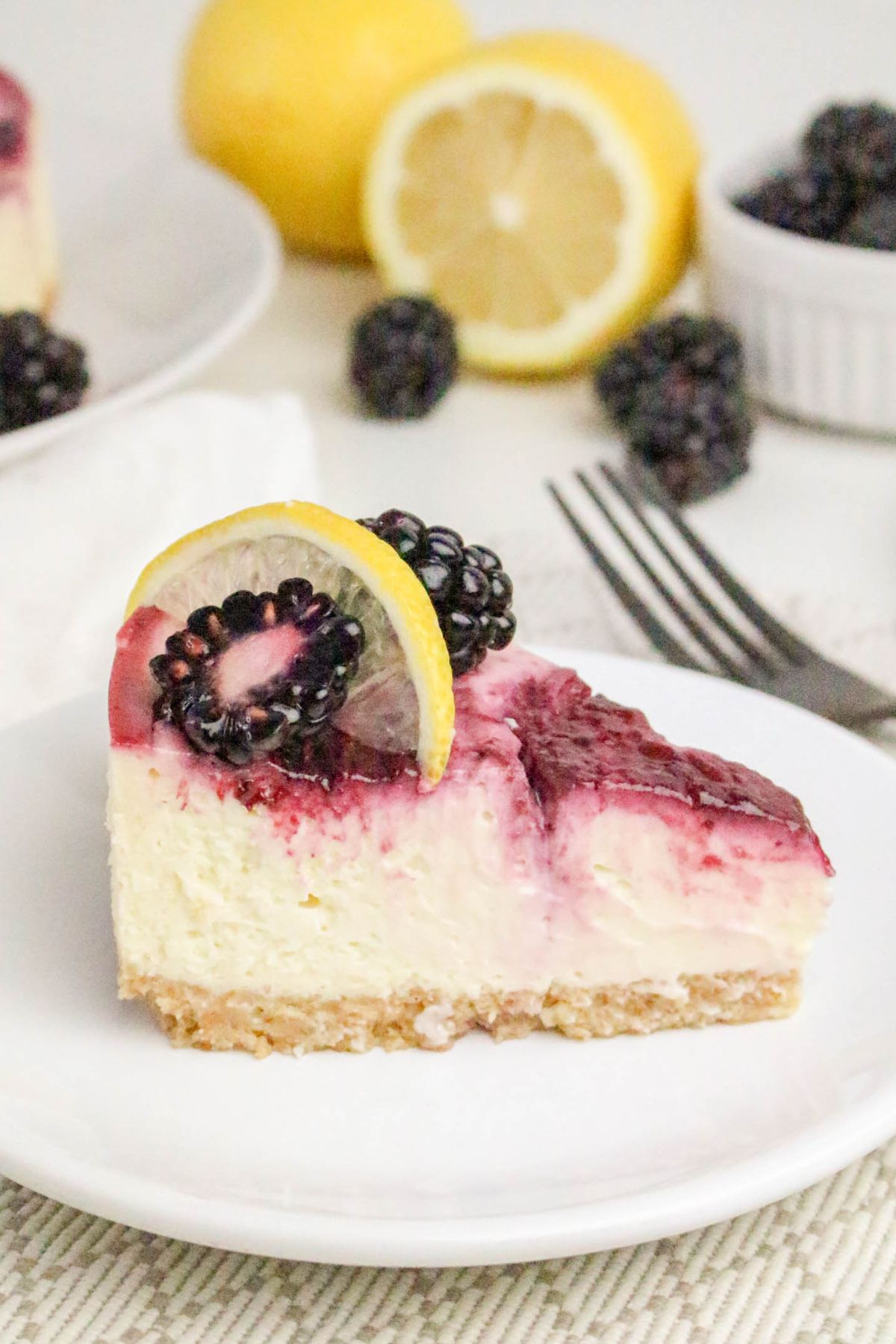 A slice of cheesecake topped with blackberry sauce, fresh blackberries, and a lemon slice, served on a white plate with a fork beside it.