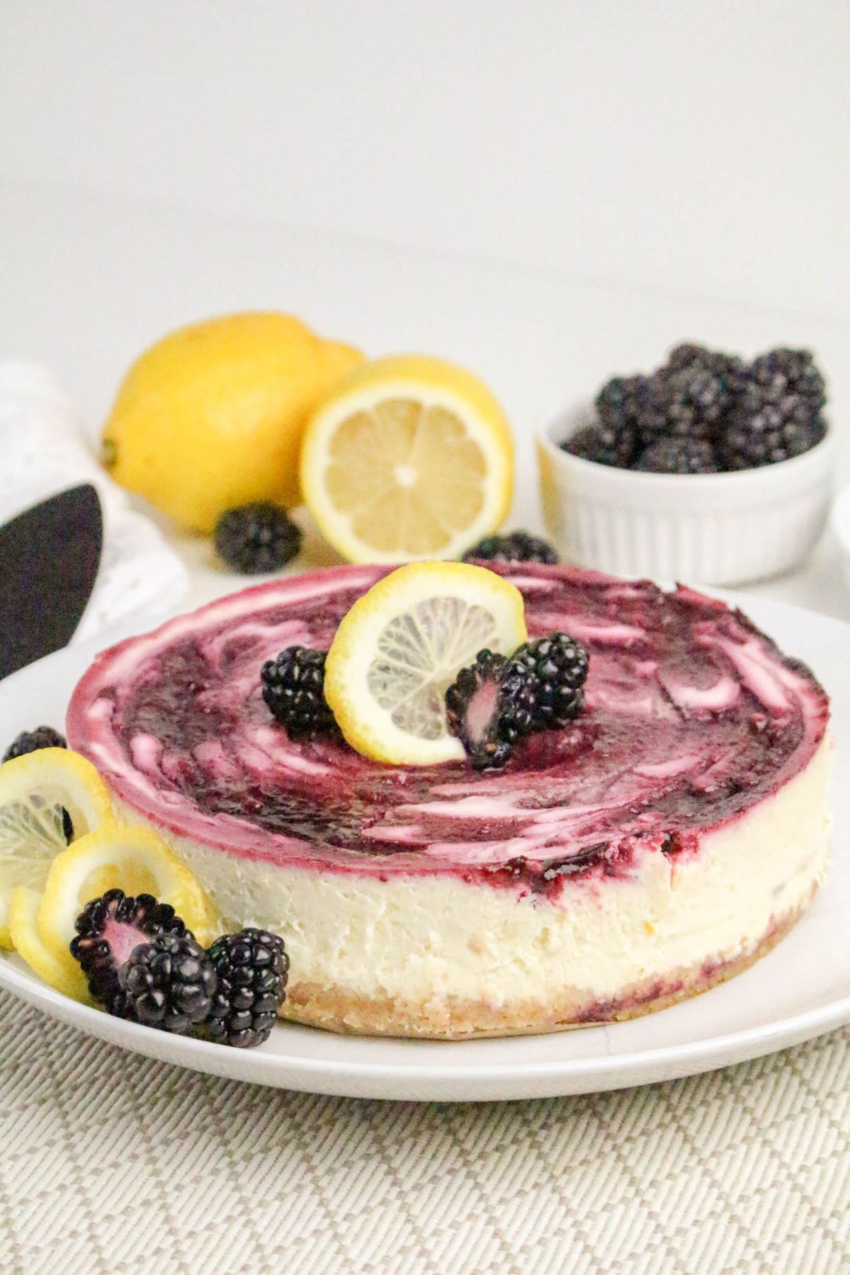 A cheesecake with a berry swirl topping, garnished with lemon slices and blackberries, sits on a plate with lemons and a bowl of blackberries in the background.