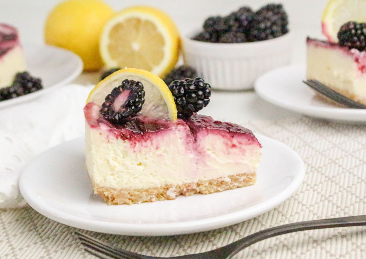 A slice of cheesecake topped with blackberry sauce, fresh blackberries, and a lemon slice sits on a white plate; lemons and blackberries are in the background.