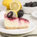 A slice of cheesecake topped with blackberry sauce, fresh blackberries, and a lemon slice on a white plate, with whole blackberries and a halved lemon in the background.