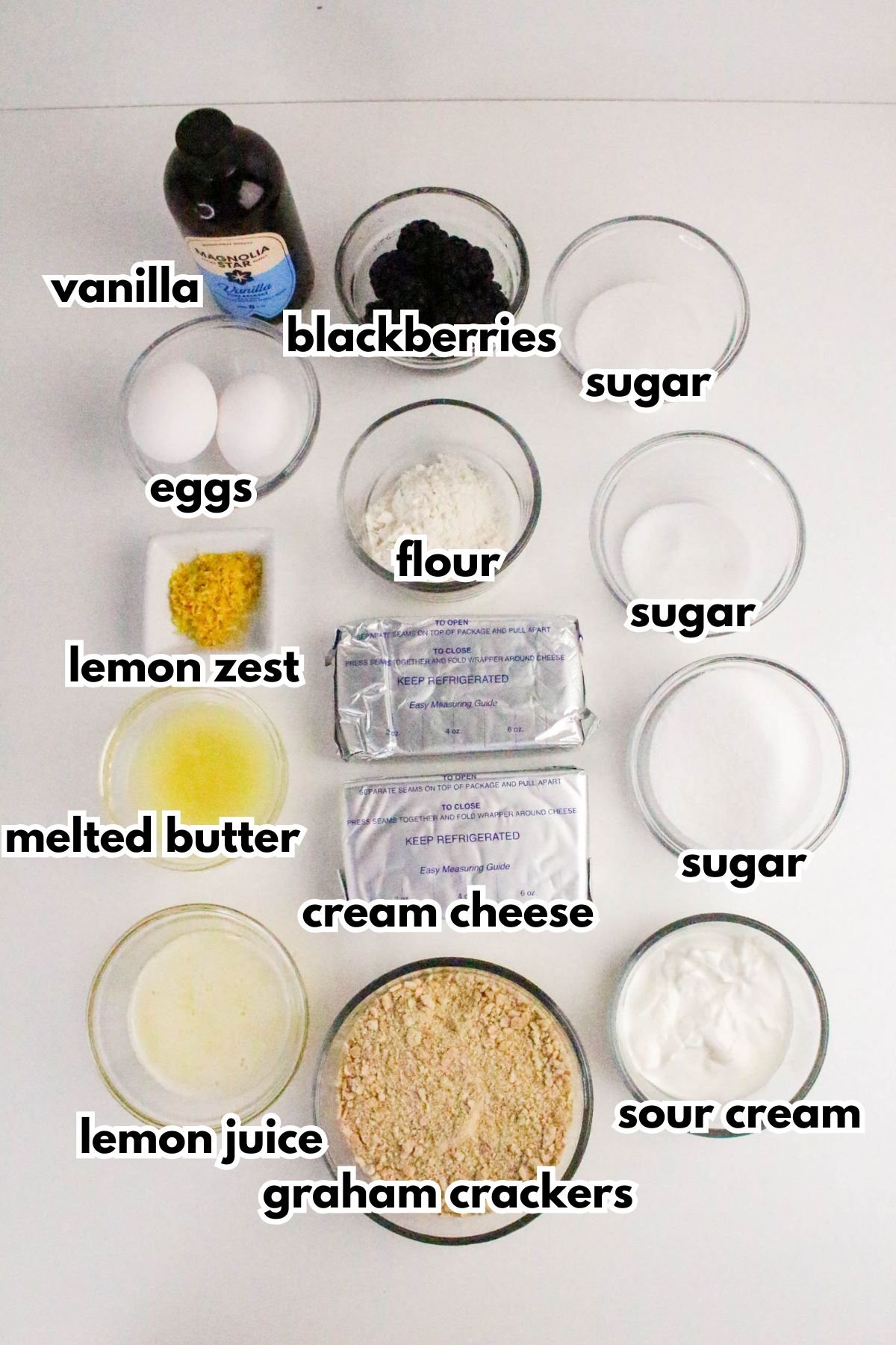 Ingredients for a blackberry cheesecake are arranged in bowls, including vanilla, blackberries, eggs, lemon zest, flour, sugar, melted butter, cream cheese, sour cream, lemon juice, and graham crackers.