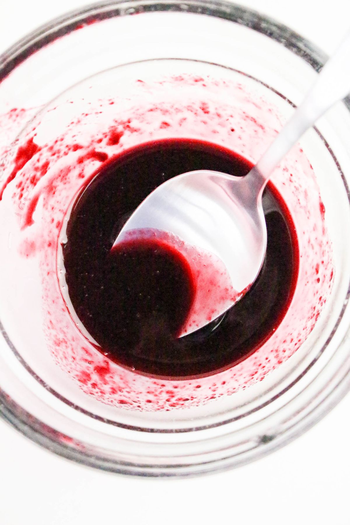 A spoon rests in a glass bowl containing a thick, dark red liquid with some residue on the sides.