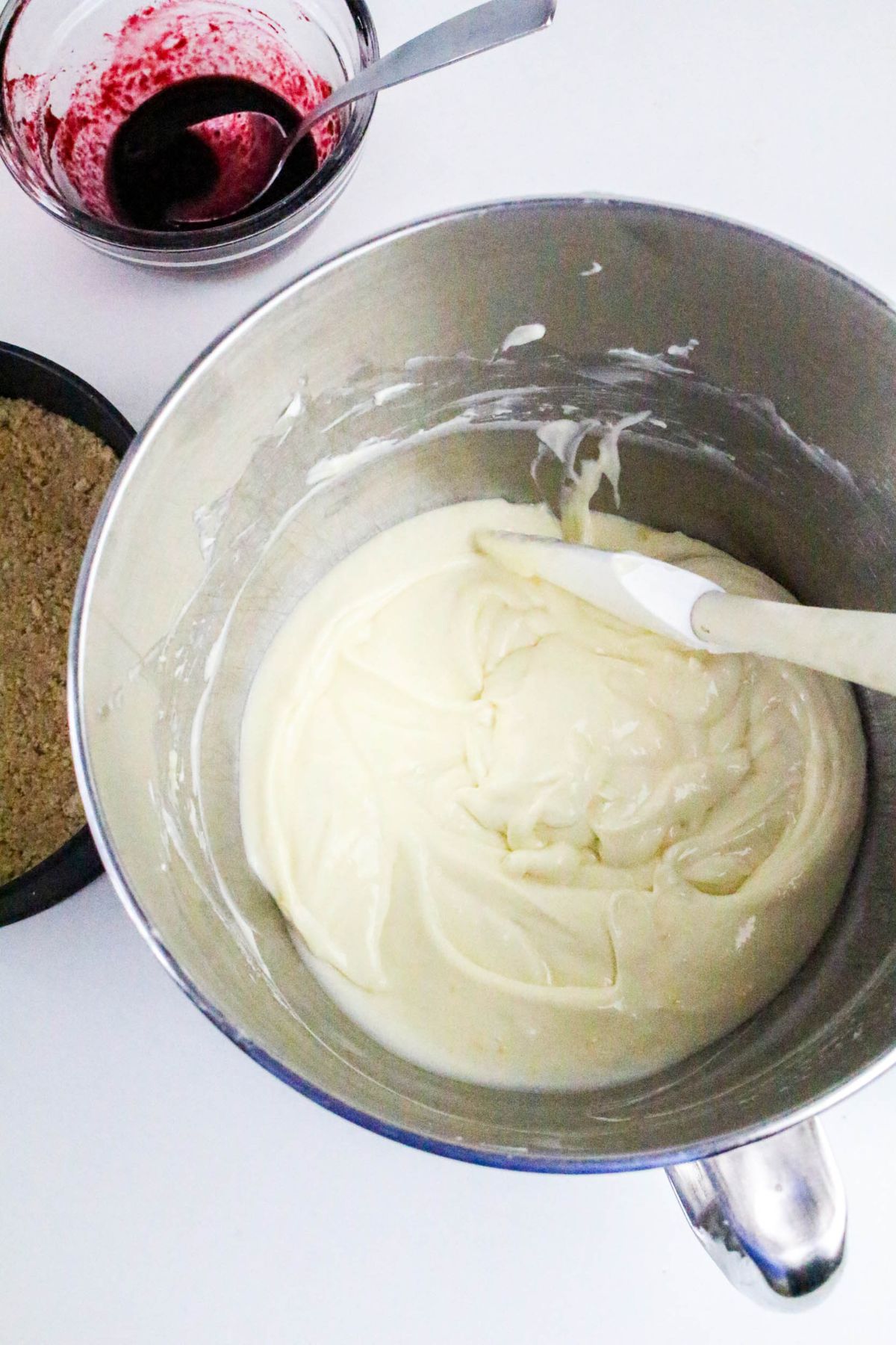 A metal mixing bowl with cheesecake batter and a spatula, a bowl of berry sauce, and a pan with crust on a white surface.