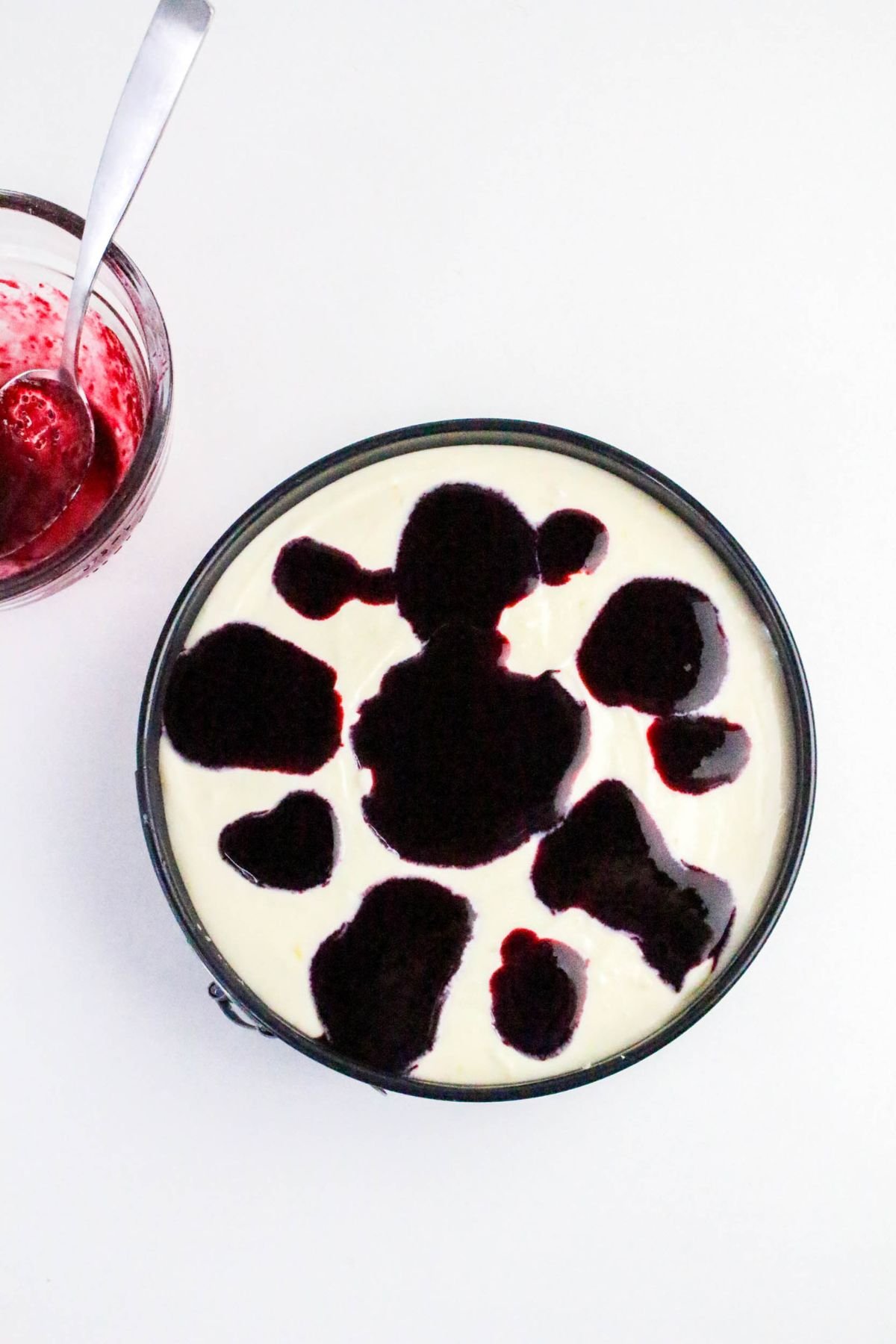 A round cheesecake with uneven blobs of dark berry sauce on top, next to a jar of berry sauce with a spoon, all on a white background.