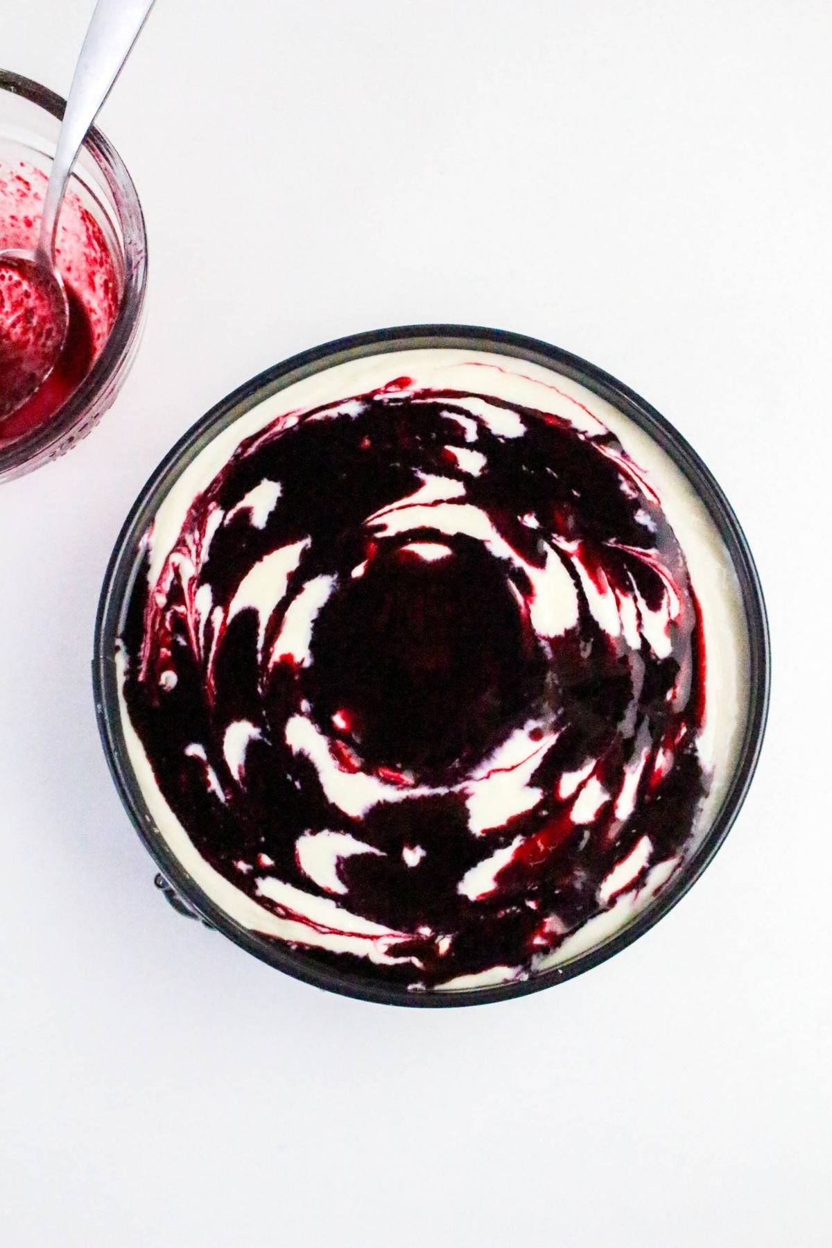 A round cheesecake with a dark red berry swirl pattern on top, next to a small bowl containing more berry sauce and a spoon.