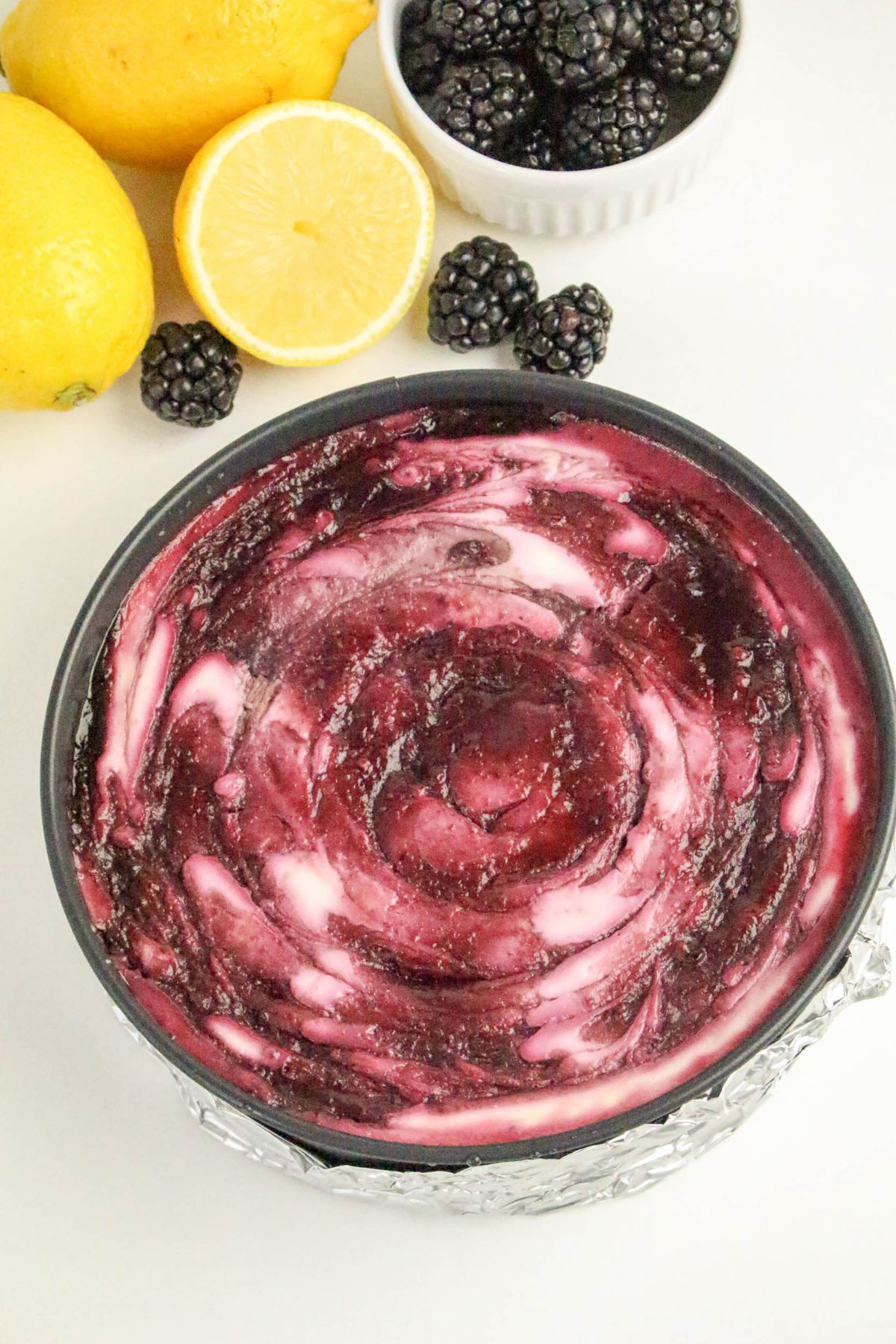 A cheesecake with a dark purple blackberry swirl on top, surrounded by fresh blackberries and lemons on a white surface.