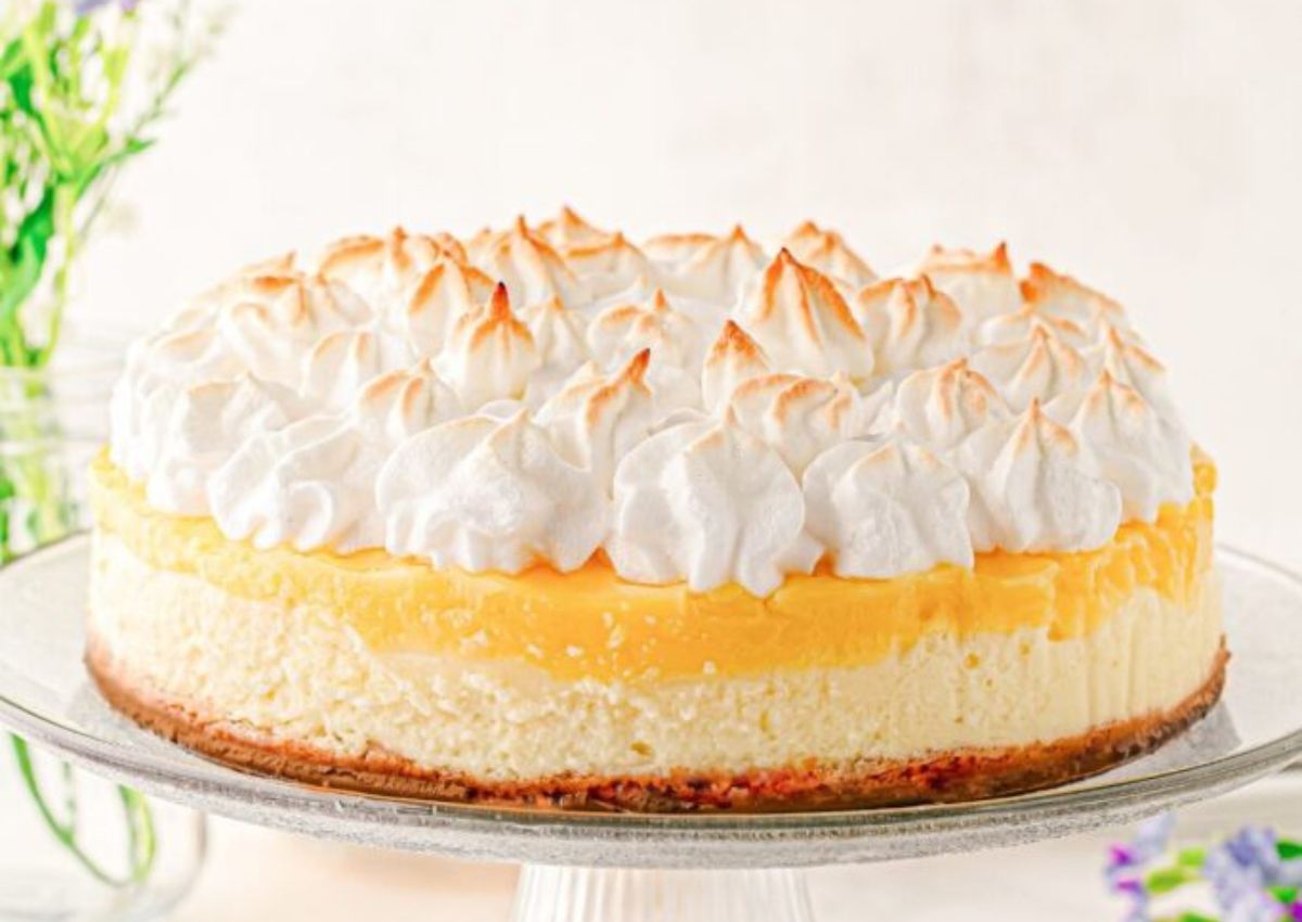 A lemon meringue cheesecake on a glass cake stand, featuring a browned meringue topping, yellow lemon layer, creamy cheesecake filling, and a brown crust.
