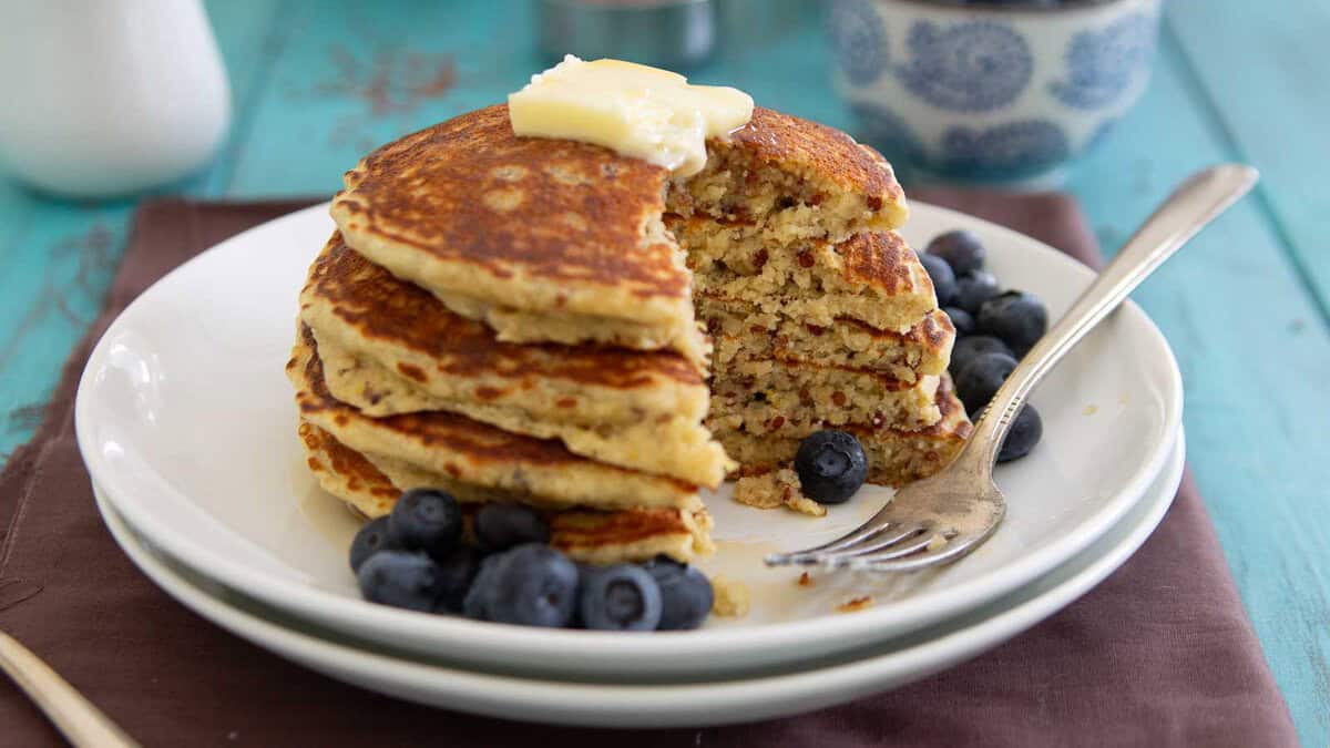 A stack of pancakes with a pat of butter on top, partially eaten, served with fresh blueberries on a white plate with a fork.