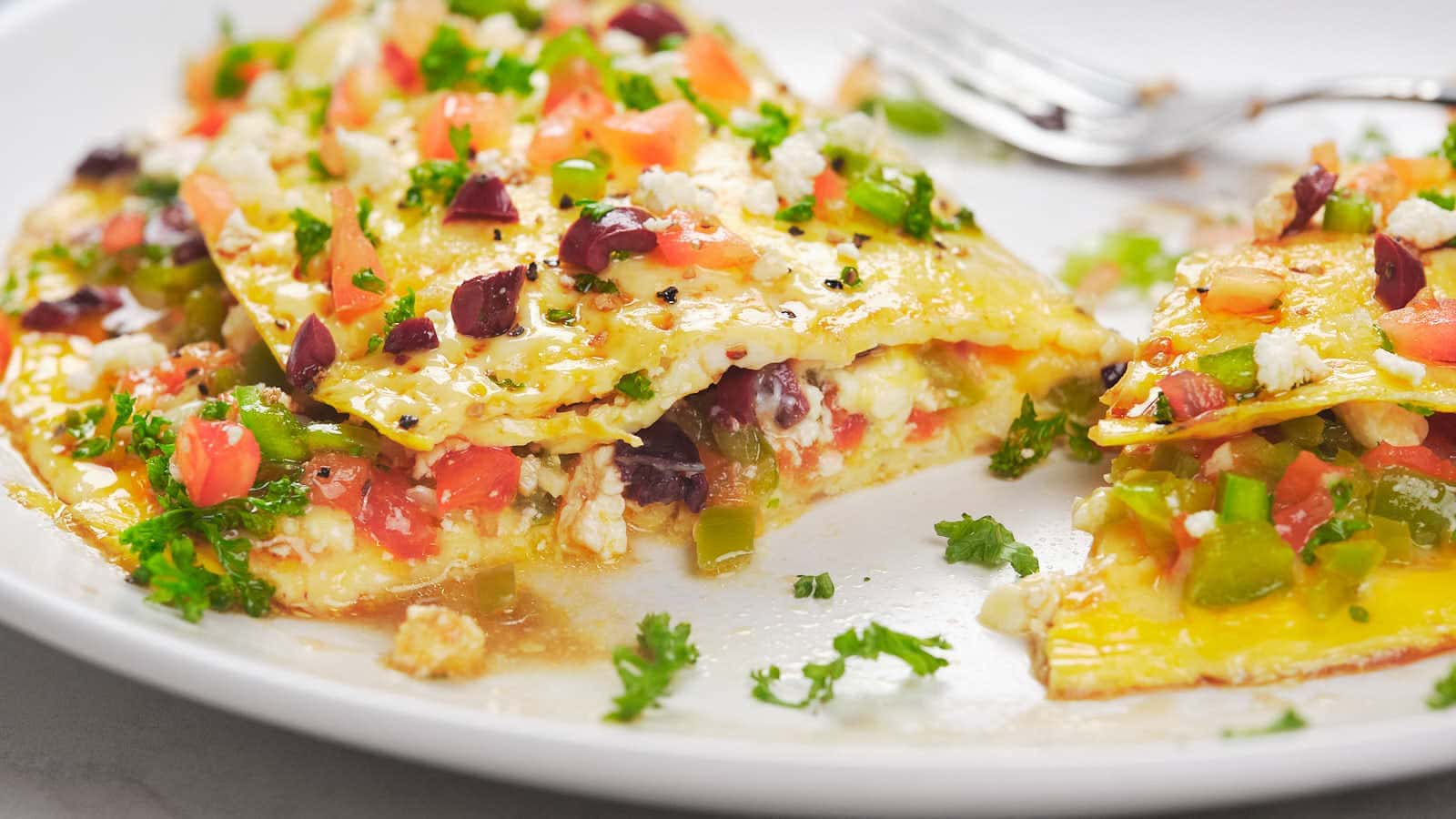 A close-up of a plate with pieces of lasagna topped with chopped tomatoes, olives, green peppers, herbs, and crumbled cheese, with a fork in the background.