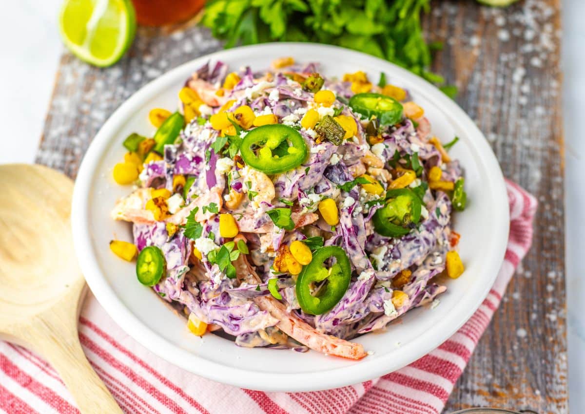 A plate of colorful salad with shredded cabbage, corn, sliced jalapeรฑos, chopped herbs, and creamy dressing, served on a white dish with a wooden spoon nearby.