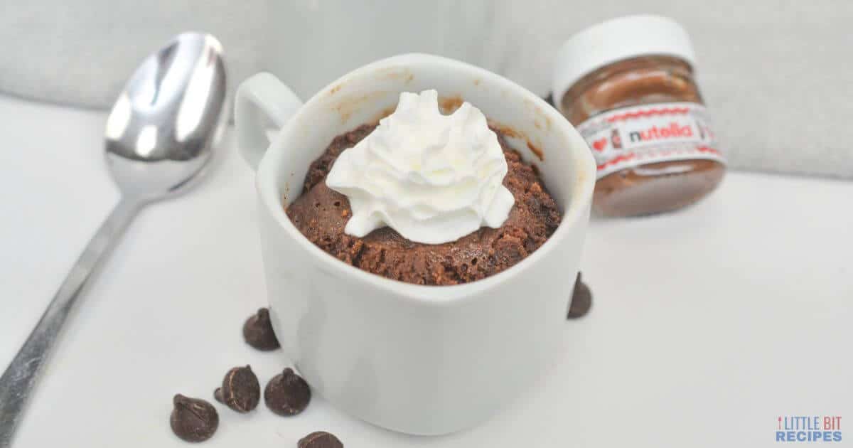 A chocolate mug cake topped with whipped cream sits beside a spoon, chocolate chips, and a small jar of Nutella on a white surface.