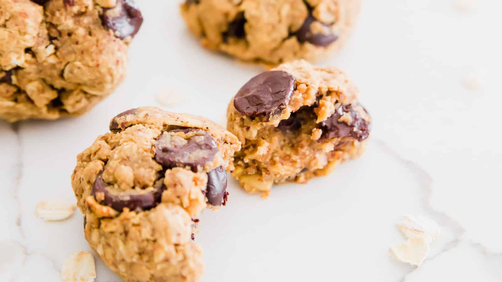 Close-up of oatmeal chocolate chip cookies on a white surface, with one cookie broken in half to show the inside texture.