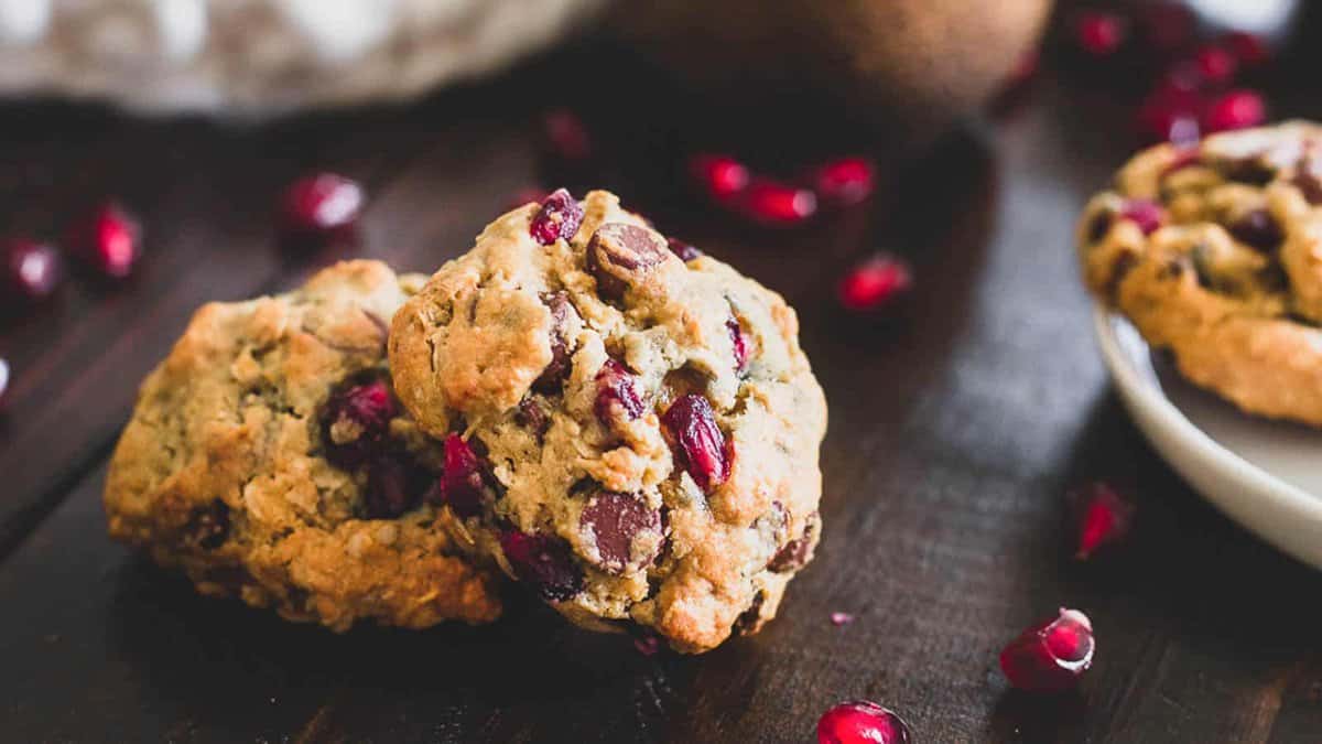 Two chocolate chip cookies with pomegranate seeds on a dark wooden surface, with scattered pomegranate seeds nearby.