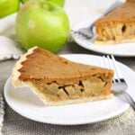A slice of apple pie with a brown, spiced filling on a white plate with a fork; whole green apples are visible in the background.