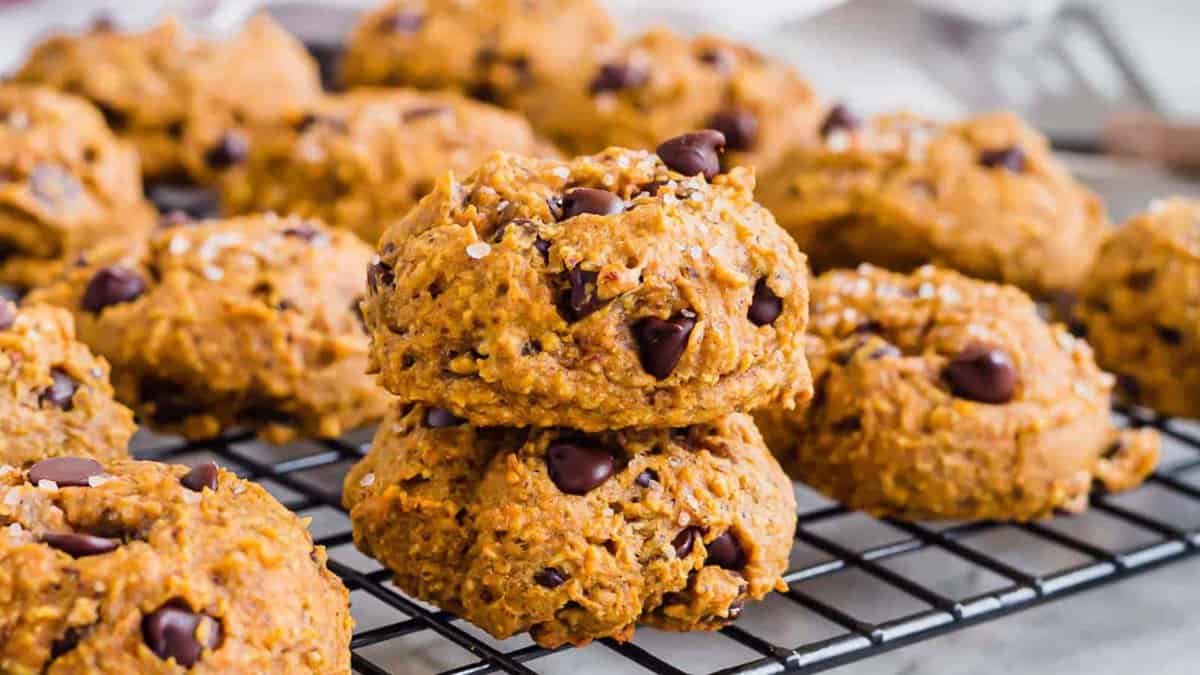 A cooling rack holds several chocolate chip cookies, some stacked, with a golden-brown, soft texture and visible chocolate chips.