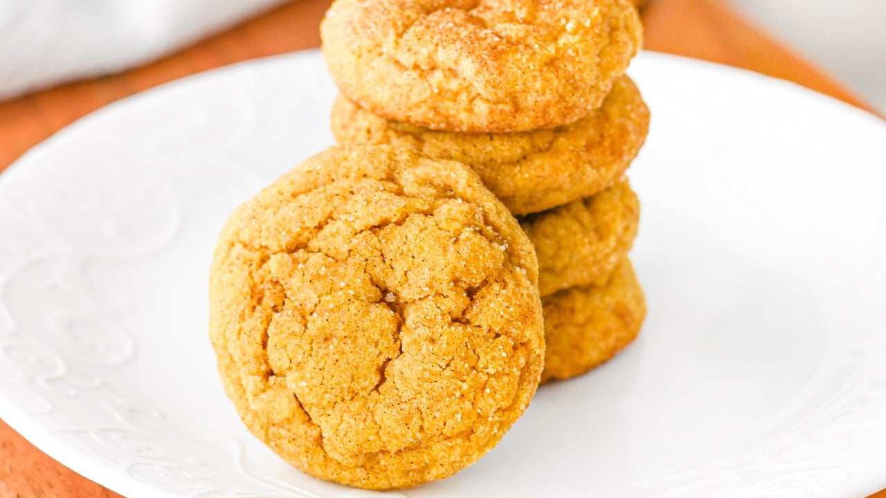 A stack of golden-brown cookies is arranged on a white plate, with one cookie leaning against the stack in the foreground.