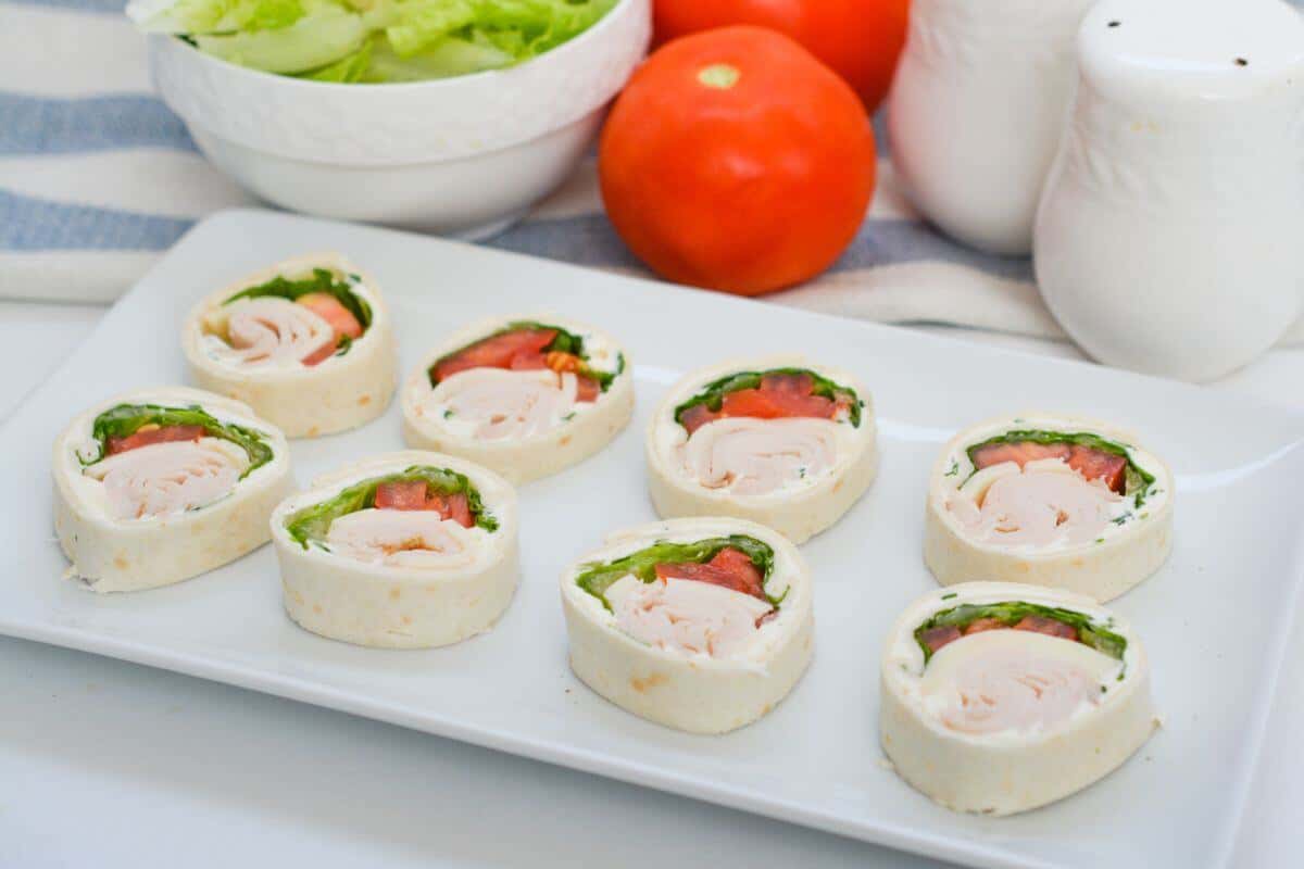 A white plate with eight turkey, lettuce, and tomato pinwheel sandwiches, with fresh tomatoes, a bowl of lettuce, and salt and pepper shakers in the background.