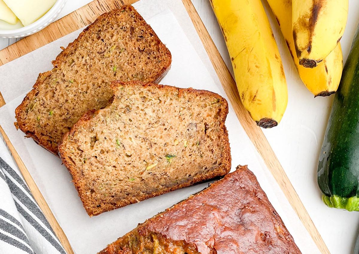 Two slices of banana zucchini bread on parchment paper beside a loaf, ripe bananas, and a zucchini on a wooden board.