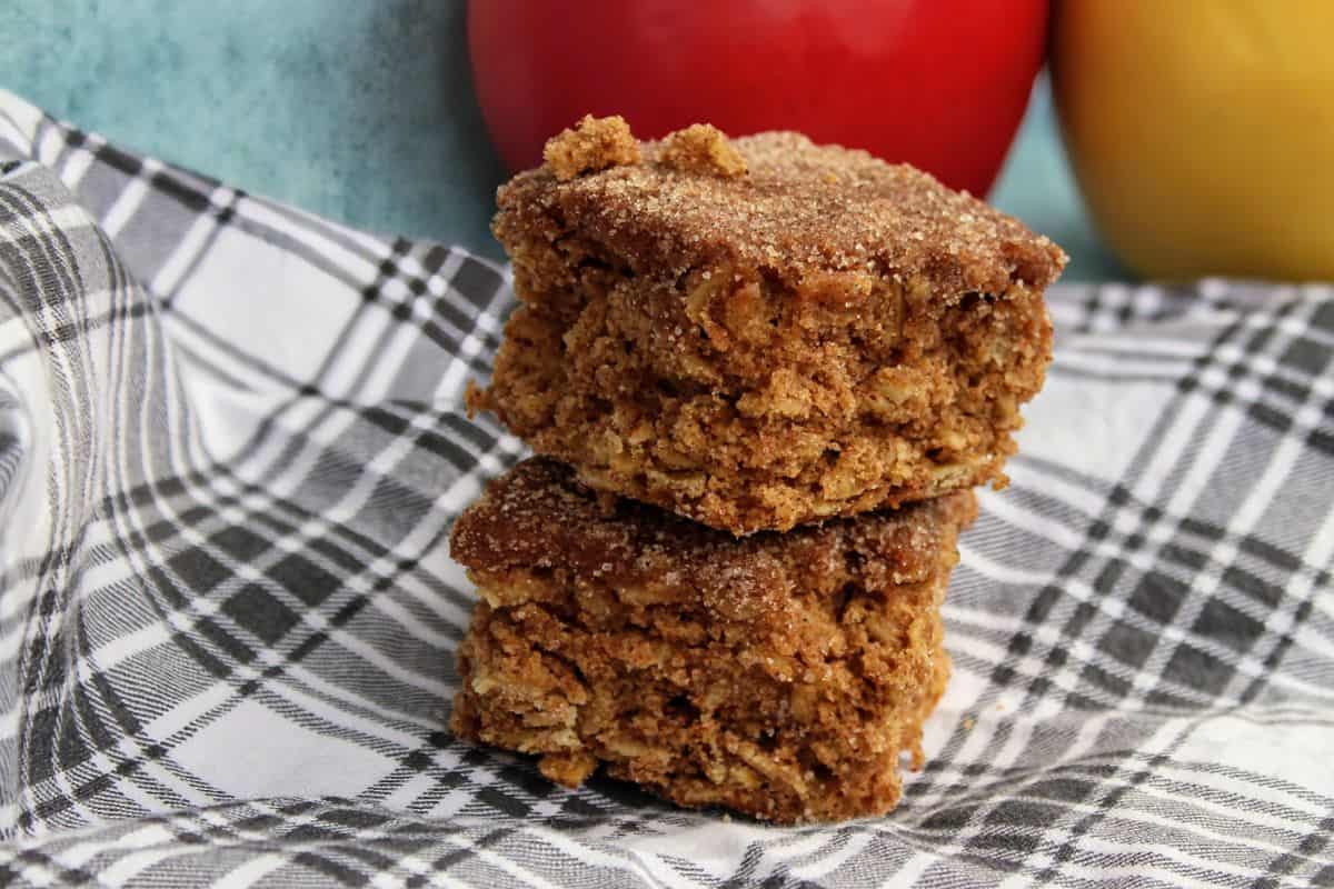 Two pieces of spiced cake bars stacked on a plaid cloth, with a red and a yellow apple in the background.