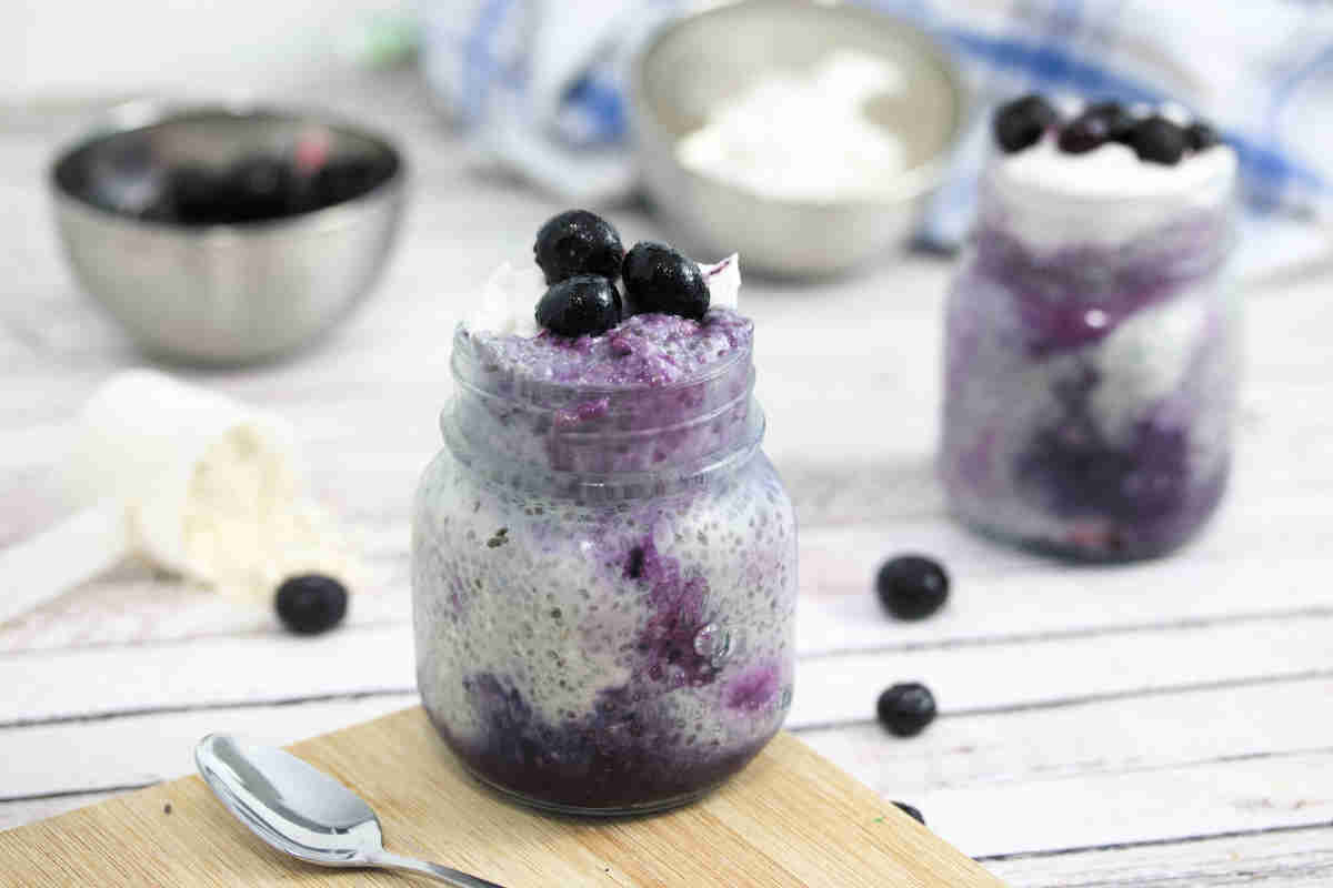 Blueberry chia pudding in a mason jar topped with coconut cream and blueberries on a wooden cutting board with blueberries and protein powder around it.