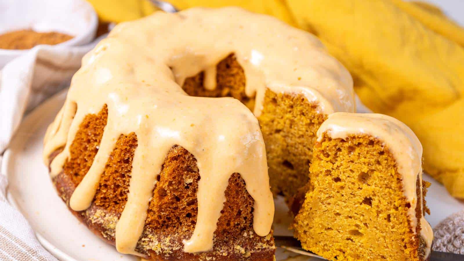 A bundt cake with creamy frosting drizzled on top, with one slice cut and slightly separated from the whole cake on a white plate.