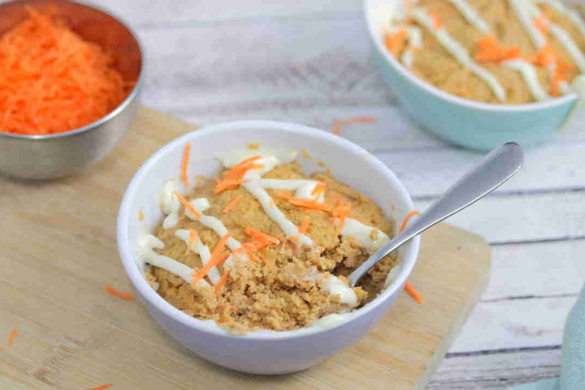 Carrot mug cake in a white ramekin with a spoon in the ramekin and cream cheese frosting on top. Ramekin on a cutting board with carrot behind it.