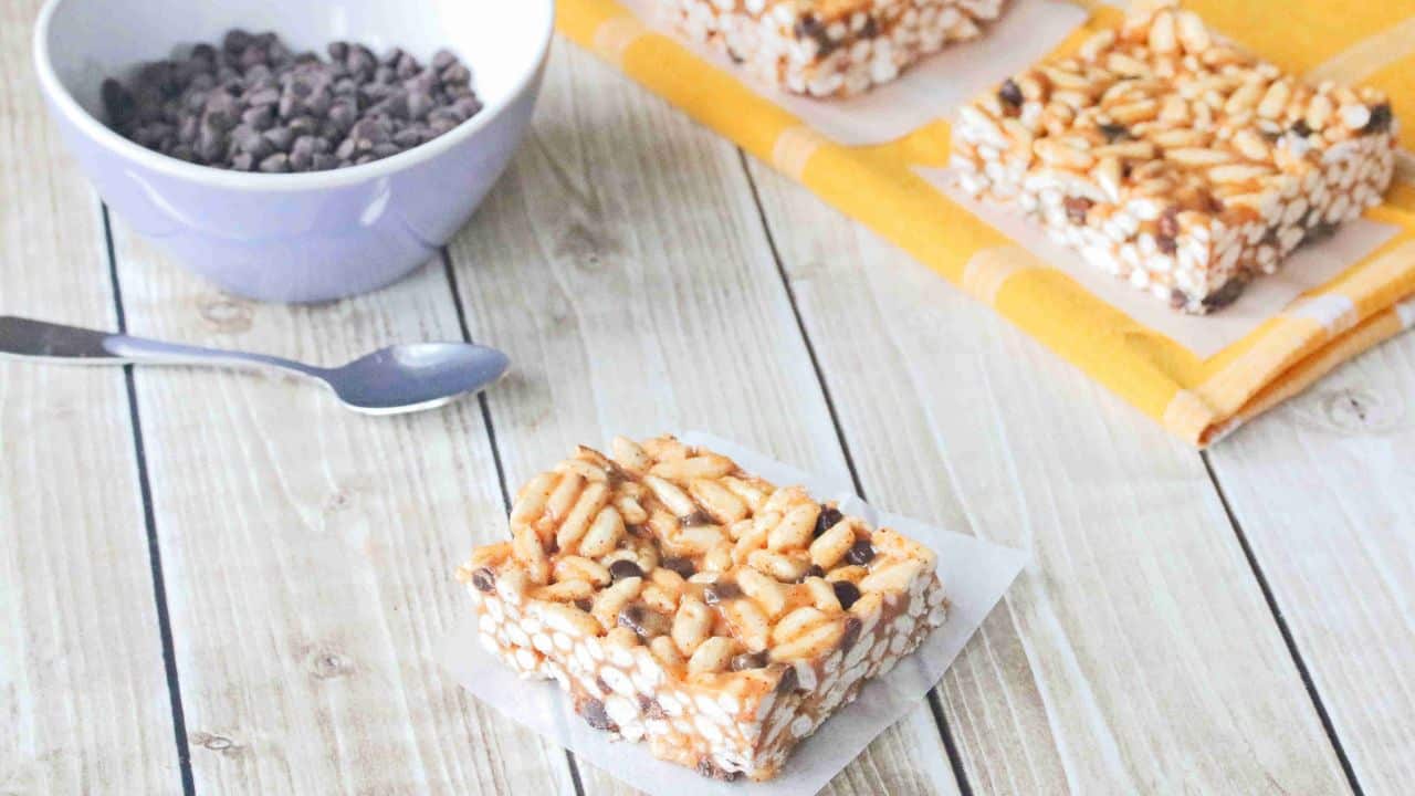 Gluten-free rice krispie square on parchment paper with more squares and a bowl of chocolate chips behind it.